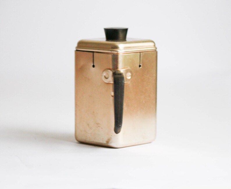 K&ouml;nnte beinhalten: Ein goldfarbener Metallbeh&auml;lter mit einem schwarzen Plastikdeckel und einem schwarzen Metallgriff. Der Beh&auml;lter hat eine rechteckige Form und ist zum Aufbewahren von Lebensmitteln gedacht.