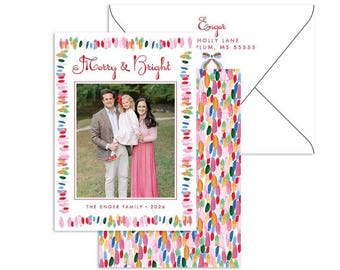 May include: A rectangular holiday card and matching envelope featuring a colorful painted brushstroke border. The card displays a photograph of three people, the text Merry & Bright, and The Enger Family 2026. The envelope shows the Enger return address.