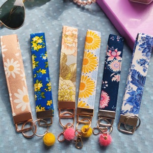 Könnte beinhalten: Sechs Schlüsselanhänger mit Stoffarmband und Blumenmustern in verschiedenen Farben, darunter Gänseblümchen, Sonnenblumen und blaue Blumenmuster. Jeder Schlüsselanhänger hat einen goldfarbenen Metallverschluss und -ring, einige mit bunten Pompons.