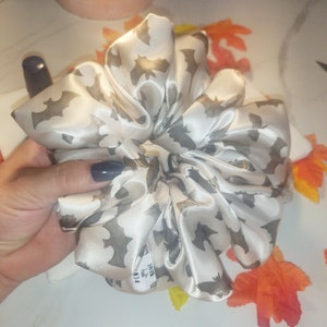 Puede incluir: Un scrunchie de satén blanco con estampado de murciélagos. El scrunchie tiene una pequeña etiqueta que dice "PCMY" y "BIG"