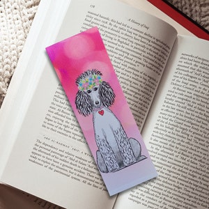 May include: A pink and purple watercolor bookmark featuring a white poodle with a flower crown and a red heart on its chest. The poodle is sitting on a light pink background.