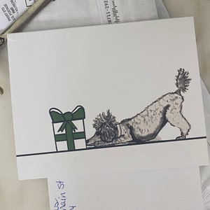 May include: A white greeting card featuring a black and white sketch of a poodle in a play bow position next to a white gift box with a dark green ribbon and bow. The illustration is set against a plain white background with a thin black line.