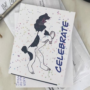 May include: A white greeting card featuring a black and white illustration of a dog standing on its hind legs with its mouth open. The card is decorated with small, multicolored confetti-like dots and the word CELEBRATE printed in blue text.