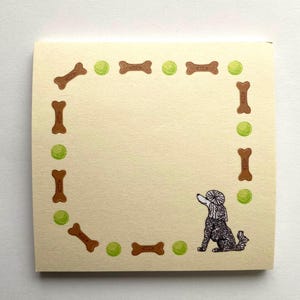 May include: A cream-colored notepad with a border of brown bones and green tennis balls. A black and white poodle with a brown hat is in the bottom right corner.
