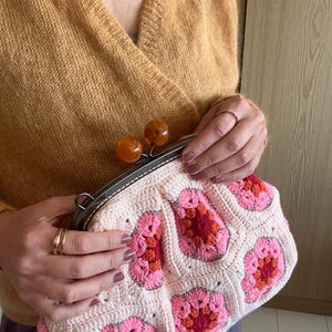 Puede incluir: Bolso de mano de ganchillo color crema con un estampado floral en tonos rosa y naranja. El bolso tiene un marco metálico con dos cierres esféricos color ámbar. La persona lleva un jersey color mostaza.