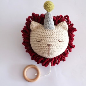 以下が含まれることがあります： 赤いたてがみと緑と灰色のパーティーハットをかぶった、かぎ針編みで作られたライオンの頭。ライオンは木製のリングが紐でつながっています。