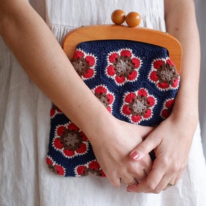 May include: A woman is holding a crocheted purse with a wooden frame. The purse is blue, red, and brown with a floral pattern.