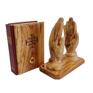Puede incluir: Sujeta libros de madera con forma de manos en oración con un diseño de cruz de Jerusalén en el libro. Los sujetalibros están hechos de madera de olivo y tienen un acabado natural. El libro es una Biblia encuadernada en cuero rojo con el título "Good News Bible" en el lomo.