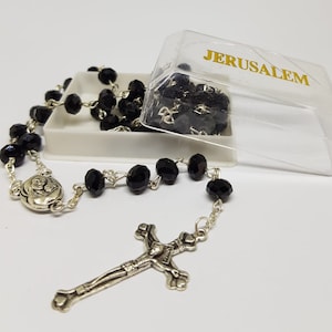May include: Black rosary beads with a silver crucifix and a silver charm featuring a mother and child. The rosary beads are in a clear plastic box with the word "JERUSALEM" printed on the top.