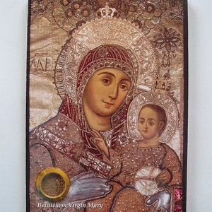 Może przedstawiać: Ikona religijna przedstawiająca Matkę Boską trzymającą Dzieciątko Jezus. Ikona jest oprawiona w ciemną drewnianą ramę i ma złote tło z misternymi detalami. Matka Boska jest ubrana w czerwoną i złotą szatę i koronę. Dzieciątko Jezus jest ubrany w białą szatę. Ikona ma mały, okrągły, złoty pojemnik przymocowany do dolnej części.
