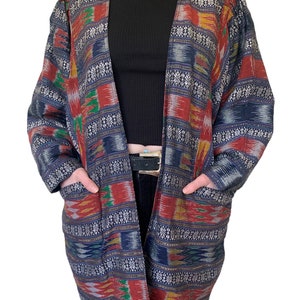 May include: A long, open-front cardigan with a colorful geometric pattern in shades of blue, red, green, gold, and silver. The cardigan has two pockets and a relaxed fit.