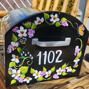 May include: Black mailbox with a white floral design and the number 1102 painted on the front. The mailbox has a silver handle and a butterfly painted on the side.