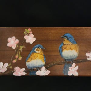 May include: A wooden panel featuring a painting of two bluebirds perched on a branch with pink cherry blossoms. The birds have blue and orange plumage. The background is a dark brown color.