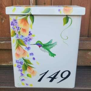 May include: White rectangular mailbox with a hand-painted design. The mailbox features a vibrant hummingbird and floral motif in shades of green, orange, purple, and yellow. The number "149" is painted in black on the front.
