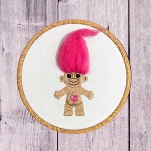 May include: Embroidered troll doll with pink hair and a pink heart on a white background in a wooden embroidery hoop.