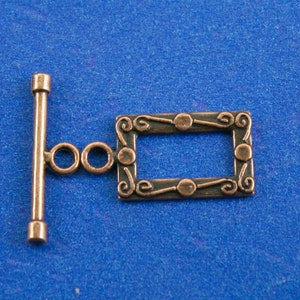 May include: A copper-toned metal toggle clasp with a decorative rectangular frame. The frame has a swirling design with small circles at each corner.