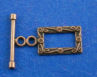 May include: A copper-toned metal toggle clasp with a decorative rectangular frame. The frame has a swirling design with small circles at each corner.