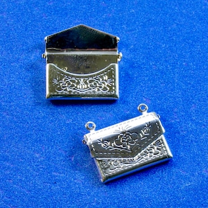 Könnte beinhalten: Zwei silberfarbene Metallanhänger in Form eines Umschlags mit Blumendesigns. Die Anhänger befinden sich auf einem blauen Hintergrund.