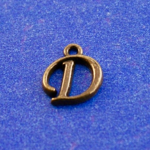 May include: A gold-colored metal charm in the shape of the letter D. The letter is stylized with a cursive font.