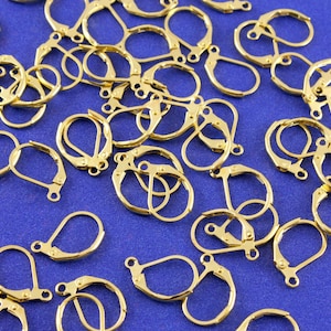 May include: A pile of gold-toned earring hooks, also known as ear wires, on a blue background. The hooks are made of metal and have a spring-loaded closure.