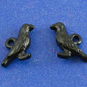 May include: Two black metal crow charms with a loop for attaching to a necklace or bracelet. The crows are facing each other and have detailed features.