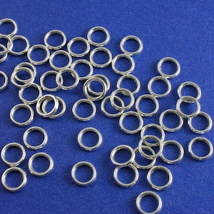 May include: A large quantity of silver jump rings on a blue background. The jump rings are small and circular, and they are all the same size.