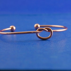 May include: A rose gold knot bangle bracelet. The bracelet is made of a thin, delicate wire and features a knot design in the center.