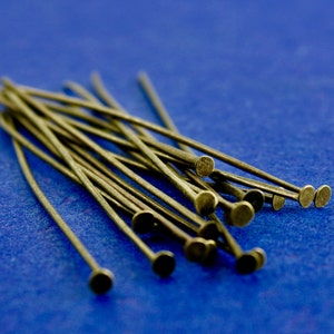 Puede incluir: Un montón de alfileres de cabeza de metal dorado con extremos redondeados. Los alfileres están dispuestos en forma de abanico sobre una superficie azul.