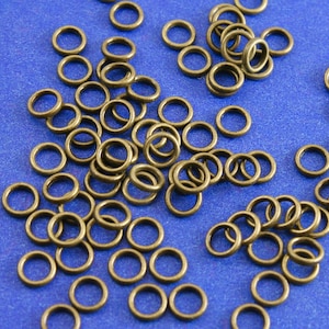 May include: A large quantity of small, gold-colored jump rings on a blue background. The jump rings are all the same size and are arranged in a random pattern.