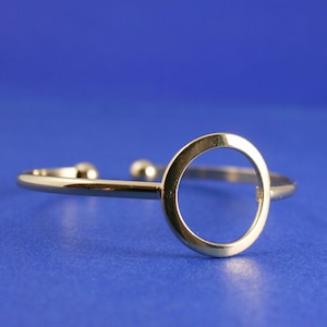 May include: A gold-toned bangle bracelet with a large, open circle design. The bracelet is made of metal and has a simple, modern style.