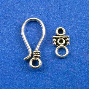 May include: Silver-toned metal jewelry clasp with a hook and a decorative loop. The hook is curved and has a twisted detail. The loop has a circular design with a small bead on each side.