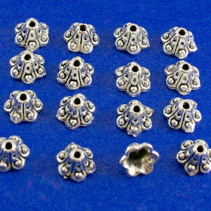 Puede incluir: Una colección de 17 tapas de cuentas de metal plateadas con un diseño floral decorativo. Las tapas están dispuestas en filas sobre un fondo azul.