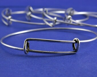 Puede incluir: Un brazalete de plata con una abertura rectangular. El brazalete está sobre un fondo azul.