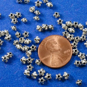 May include: A close-up of a pile of silver metal beads with a raised star design. The beads are scattered on a blue surface next to a US penny.