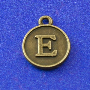 May include: A bronze-colored metal charm with the letter "E" in a serif font. The charm has a loop at the top for attaching to a necklace or bracelet.
