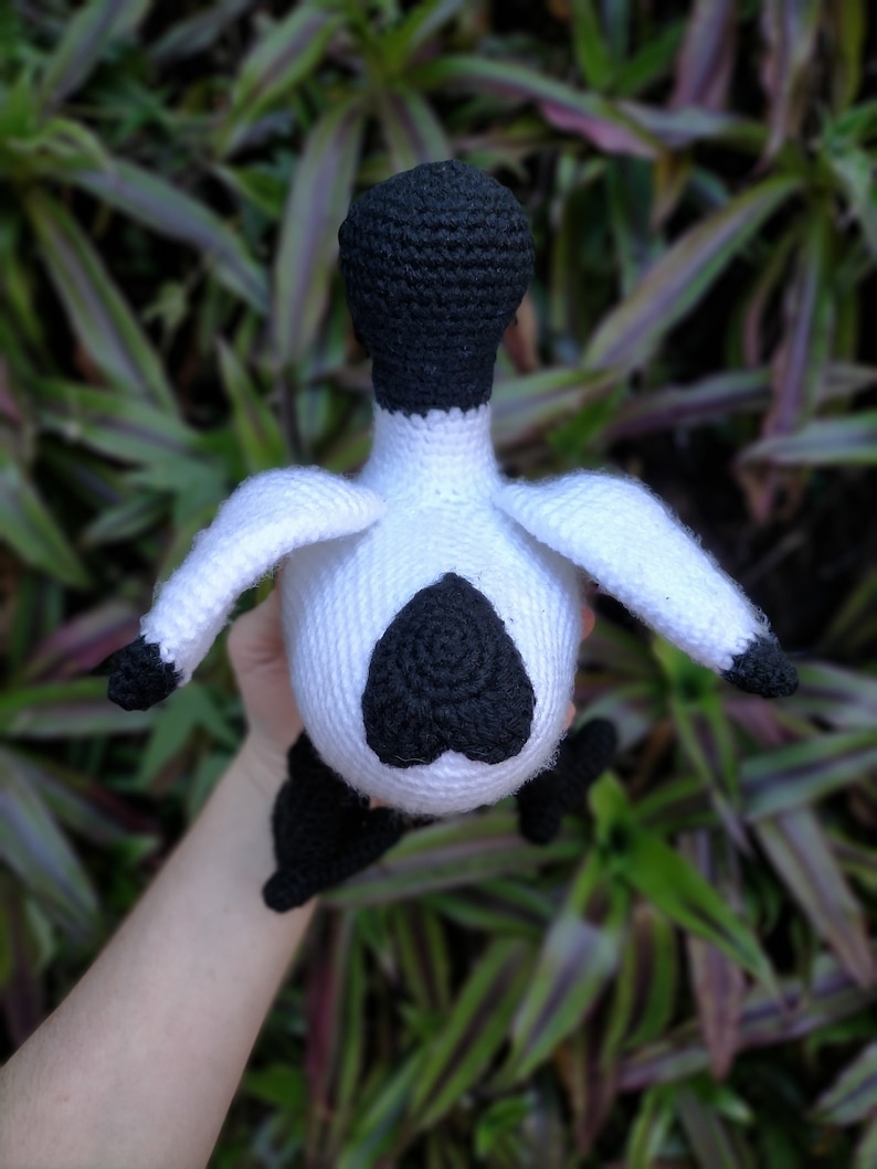 May include: A crocheted stuffed animal in the shape of a bird. The bird is white with black accents, including a black head and a black heart-shaped design on its chest.