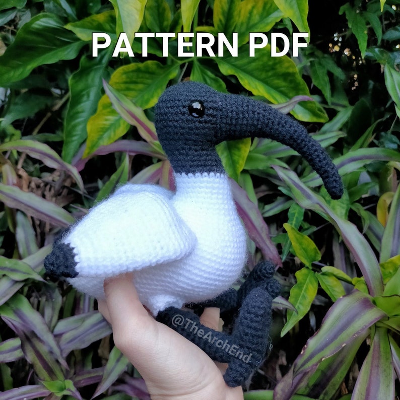 May include: Crochet pattern PDF for a white and black ibis bird amigurumi. The bird is sitting on a hand with green foliage in the background. The pattern is available for download.  @TheArchEnd
