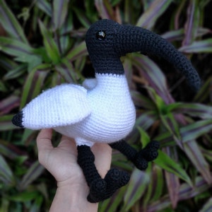 May include: A white and black crocheted stuffed bird with a long beak. The bird has black eyes and is sitting in a person's hand.