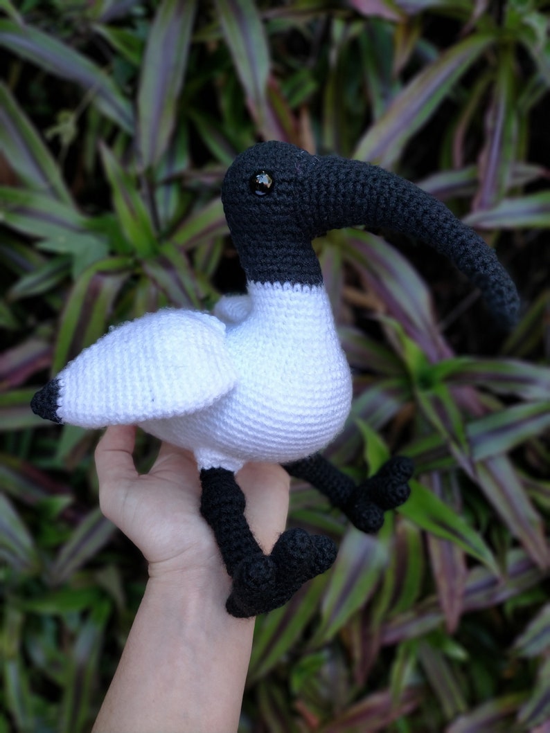 May include: A white and black crocheted stuffed bird with a long beak. The bird has black eyes and is sitting in a person's hand.