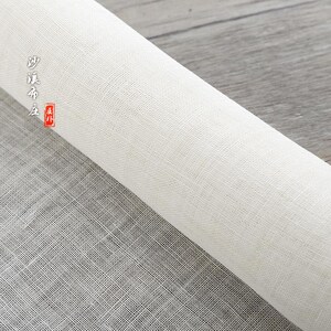Puede incluir: Un rollo de tela blanca tejida con una textura fina y uniforme. La tela est&aacute; enrollada y el extremo del rollo es visible. La tela est&aacute; etiquetada en caracteres chinos.