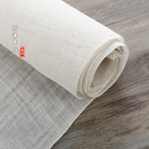 Puede incluir: Un rollo de tela blanca natural con textura tejida. La tela est&aacute; enrollada apretadamente y tiene una etiqueta en caracteres chinos.