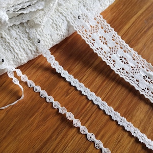 Puede incluir: Tres encajes blancos, uno con un delicado patrón floral, otro con un borde festoneado simple y el último con un diseño más intrincado. Los encajes están dispuestos sobre una superficie de madera.