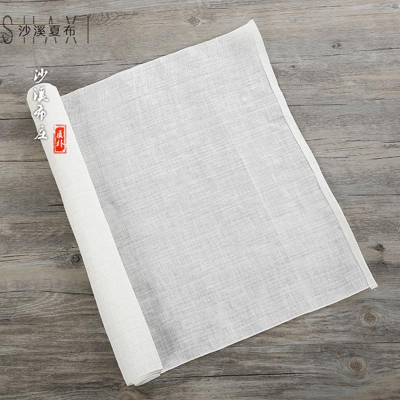 Puede incluir: Un trozo de tela blanco rectangular, enrollado sobre una superficie de madera. La tela es lisa y tiene una textura sutil. El texto "沙溪夏布" es visible en la esquina superior izquierda de la imagen.