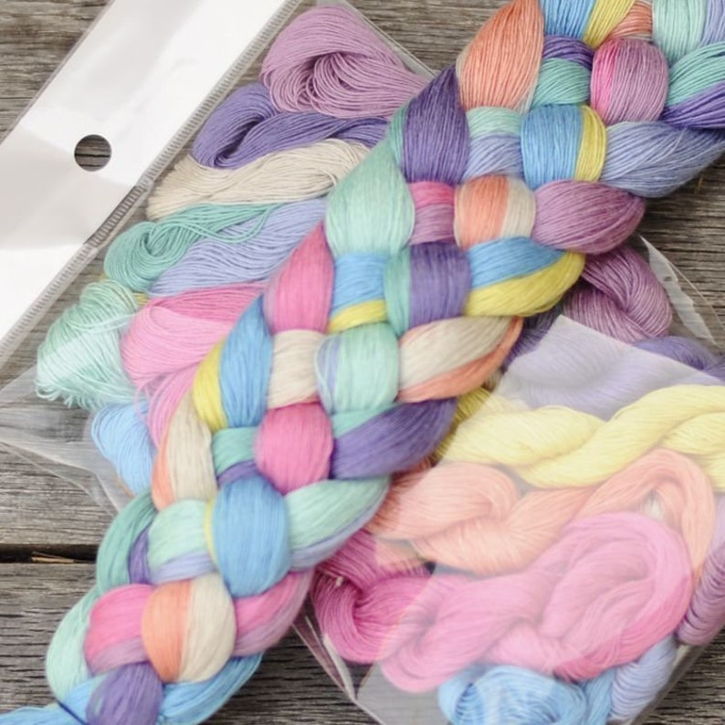 May include: Pastel embroidery floss in a clear plastic package. The floss is arranged in a braid pattern, displaying shades of pink, blue, yellow, and purple. Additional bundles of floss in various pastel hues are visible in the background.