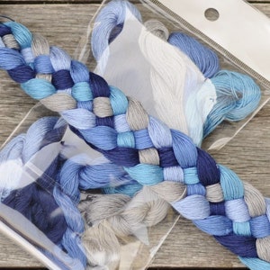 May include: A selection of embroidery floss in shades of blue and grey, presented in a clear plastic package. The floss is arranged in a decorative braid, highlighting the different colours and textures of the thread. The background is a wooden surface.