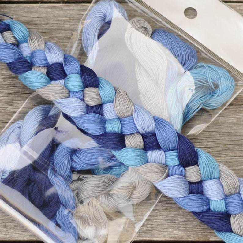 May include: A selection of embroidery floss in shades of blue and grey, presented in a clear plastic package. The floss is arranged in a decorative braid, highlighting the different colours and textures of the thread. The background is a wooden surface.