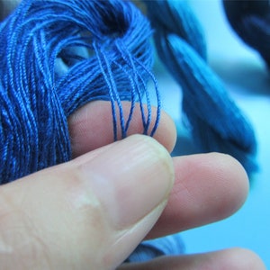 May include: Close-up of bright blue embroidery thread. A hand holds a small bunch of the thread, highlighting its fine texture and colour. Other bundles of blue thread are in the background, suggesting a craft or textile project.