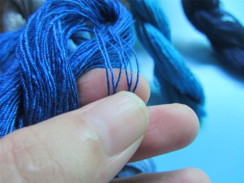 May include: Close-up of bright blue embroidery thread. A hand holds a small bunch of the thread, highlighting its fine texture and colour. Other bundles of blue thread are in the background, suggesting a craft or textile project.