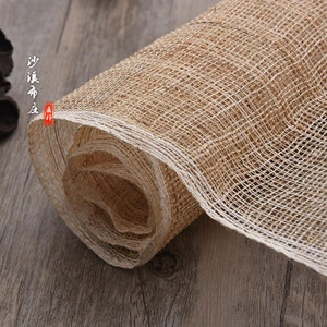 May include: A roll of natural beige and white woven fabric with a rough texture. The fabric is rolled up and has a white edge. The fabric is on a wooden surface.
