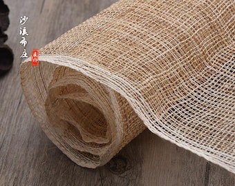 May include: A roll of natural beige and white woven fabric with a rough texture. The fabric is rolled up and has a white edge. The fabric is on a wooden surface.